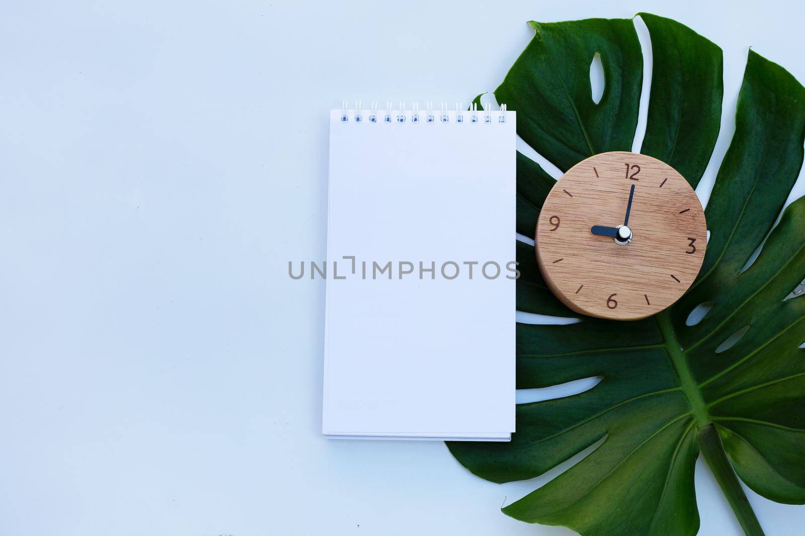 Wooden clock with notebook on green leaves on white background. Copy space by Bowonpat