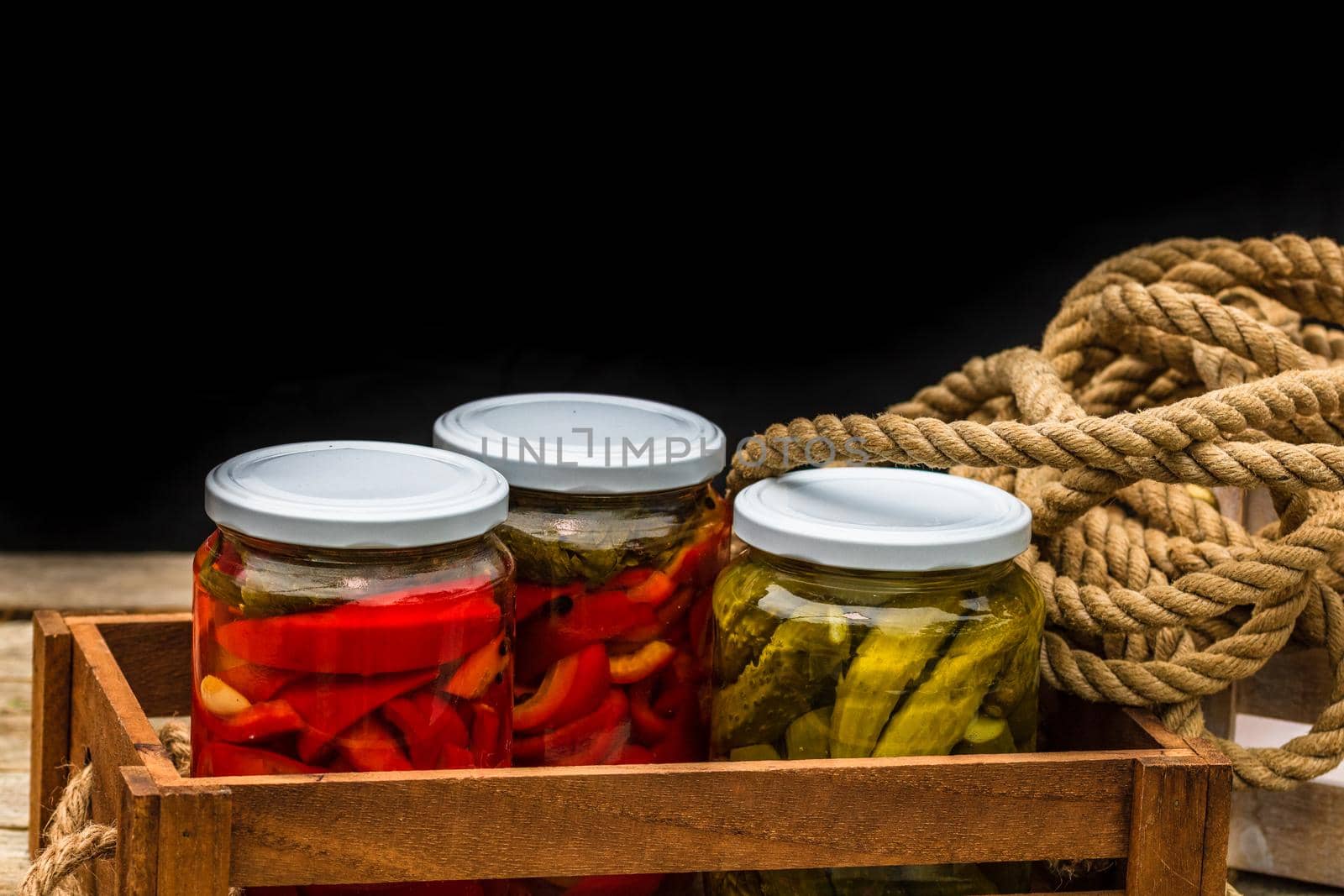 Wooden crate with glass jars with pickled red bell peppers and pickled cucumbers (pickles) isolated. Jars with variety of pickled vegetables. Preserved food concept in a rustic composition. by vladispas