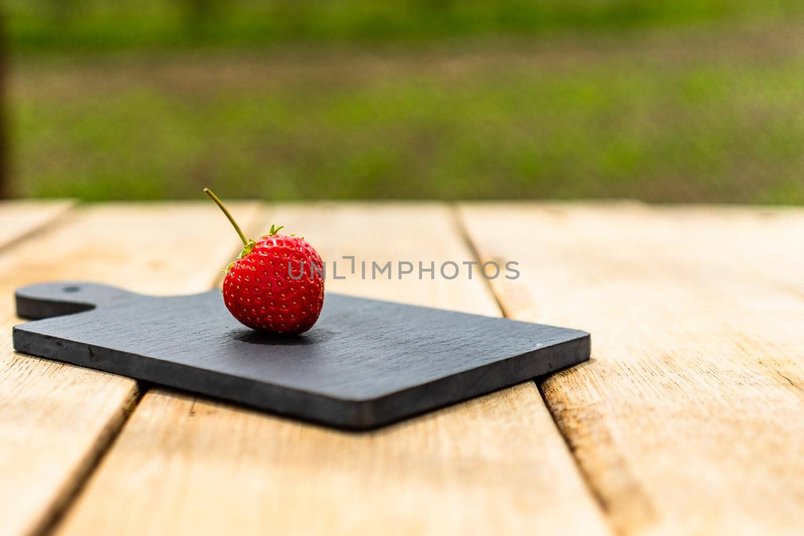 Close up of one strawberry on small black cutting board isolated outdoor on wooden table. by vladispas