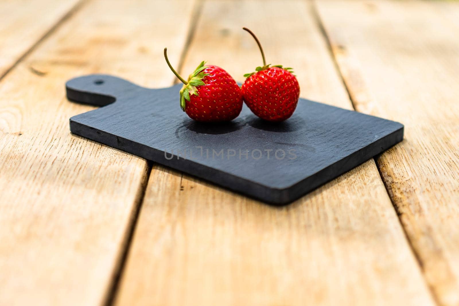 Close up of two strawberries on small black cutting board isolated outdoor on wooden table.