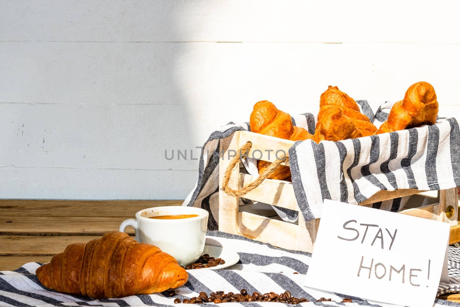 Coffee cup and buttered fresh French croissant on wooden crate. Food and breakfast concept. Morning message “stay home” on white board
