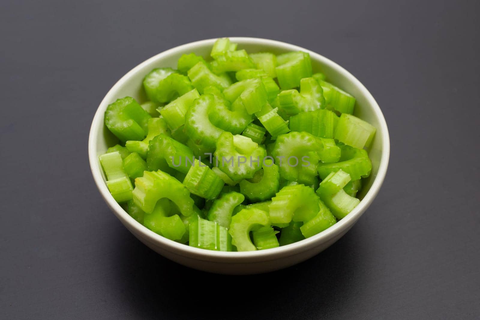 Green fresh celery pieces in white bowl on dark background. 