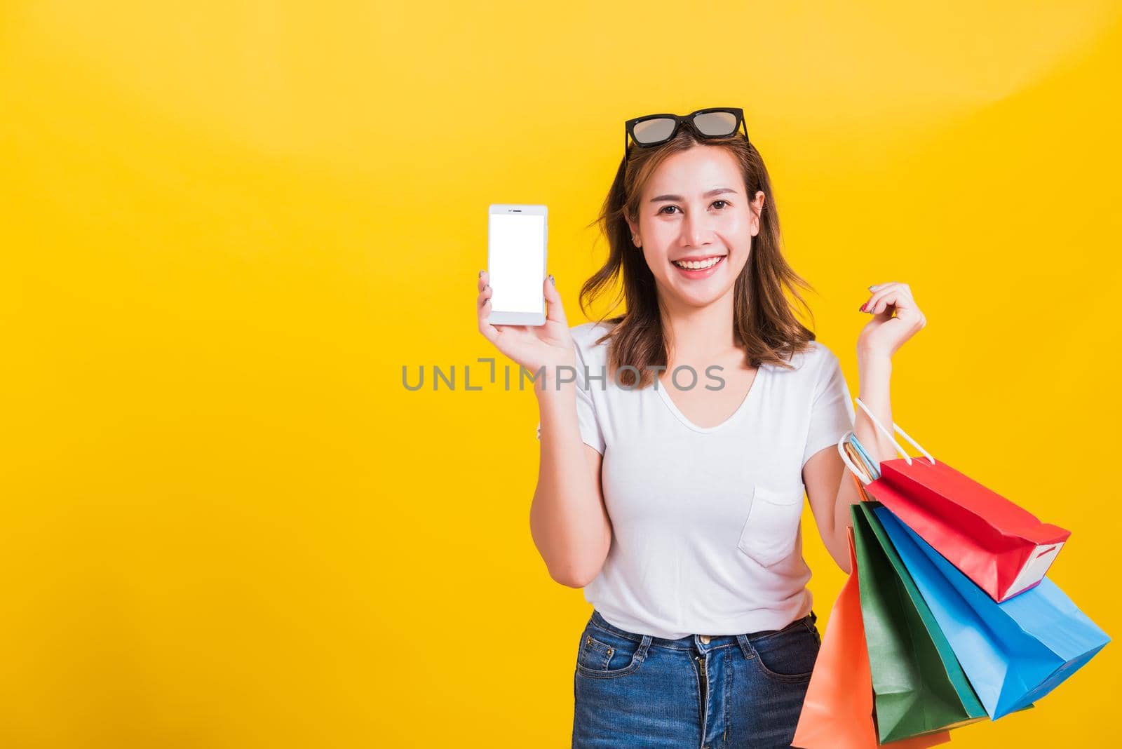 Portrait happy Thai Asian beautiful young woman smile white teeth standing wear t-shirt, She holding shopping bags and show mobile phone blank screen, studio shot on yellow background with copy space