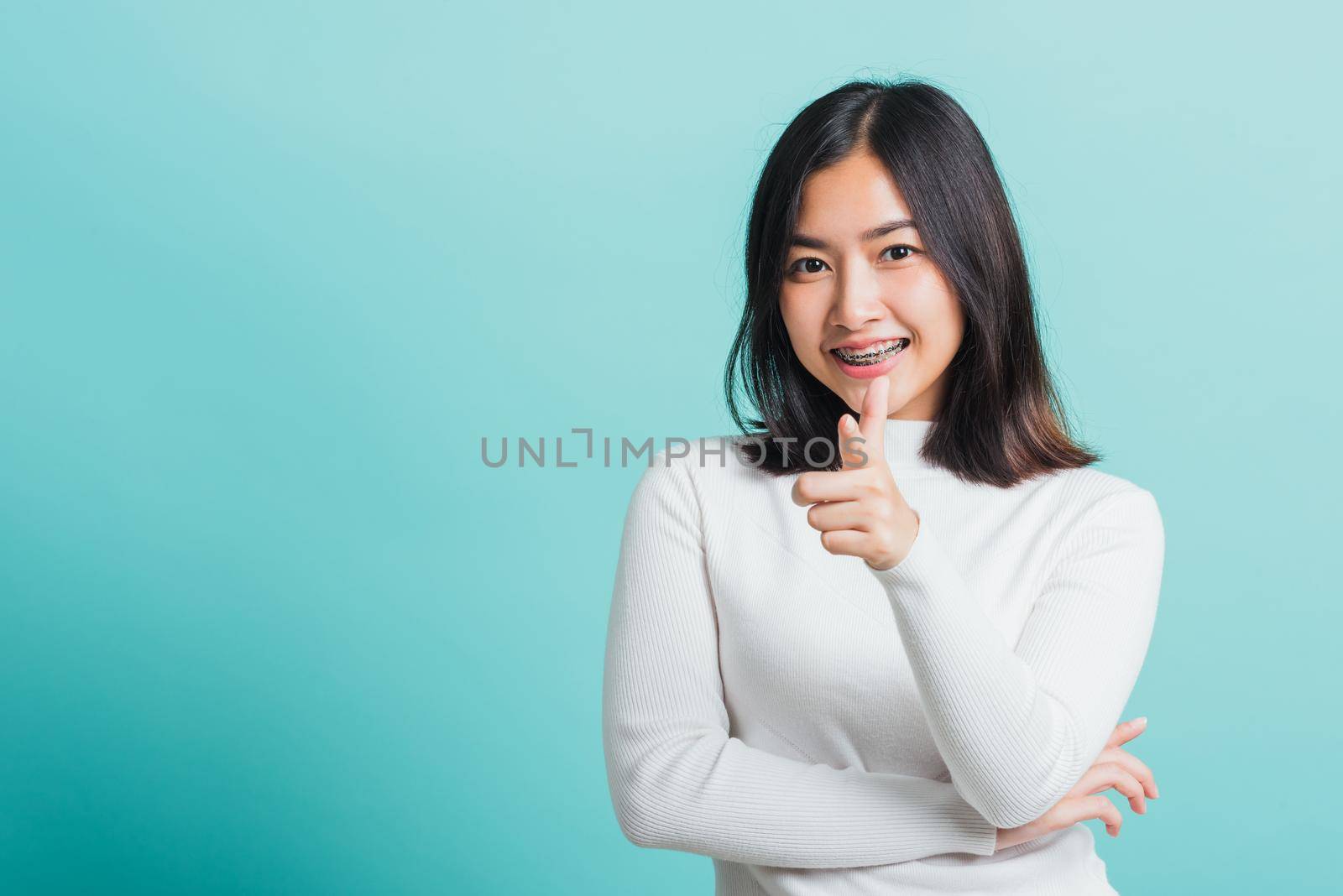 Young beautiful Asian woman smiling point finger at you with a confident expression, Portrait female pointing finger gesture towards you, studio shot isolated on a blue background