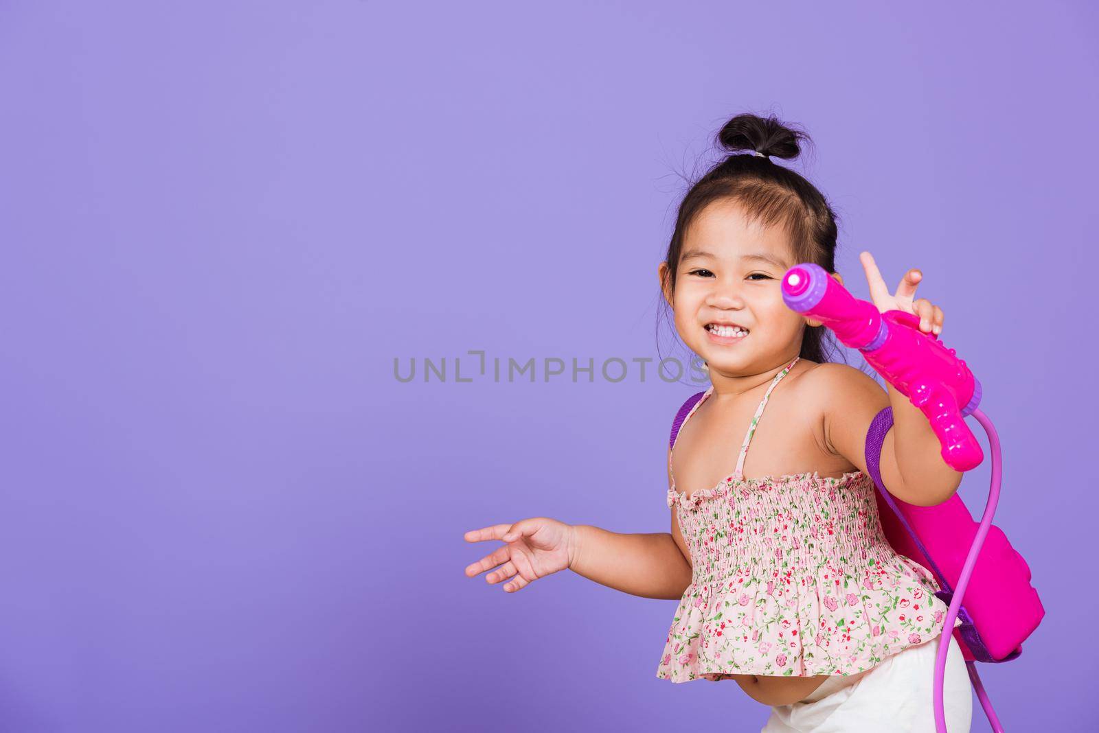 Thai child funny hold toy water pistol and smile, Happy Asian little girl holding plastic water gun, studio shot isolated on purple background, Thailand Songkran festival day national culture concept