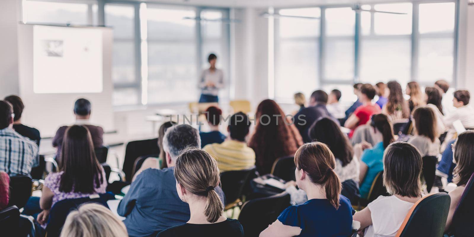 Business and entrepreneurship symposium. Speaker giving a talk at business meeting. Audience in conference hall. Rear view of unrecognized participant in audience.