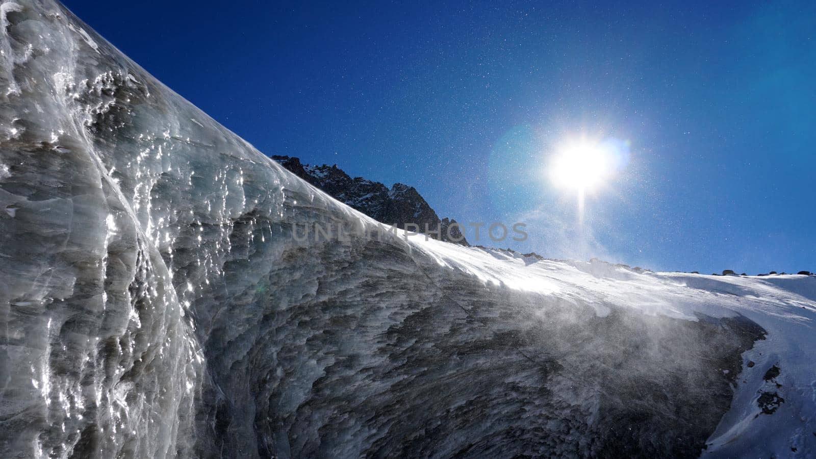 High ice wall in the mountains. The Bogdanovich Glacier. The black-and-white ice mixed. Grains of snow fly from the top of the glacier. The sun is shining. Lots of snow. Stones were frozen in places