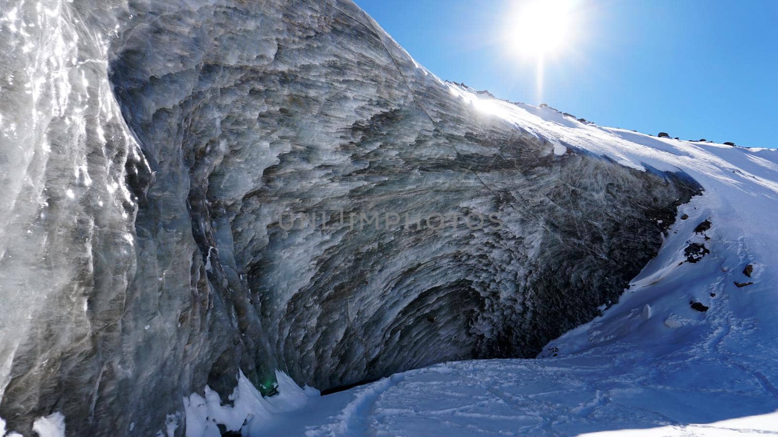 High ice wall in the mountains. The Bogdanovich Glacier. The black-and-white ice mixed. Grains of snow fly from the top of the glacier. The sun is shining. Lots of snow. Stones were frozen in places