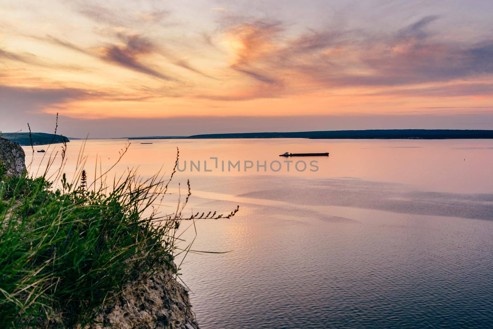 Cliff with grass and flowers with sunset on river on background