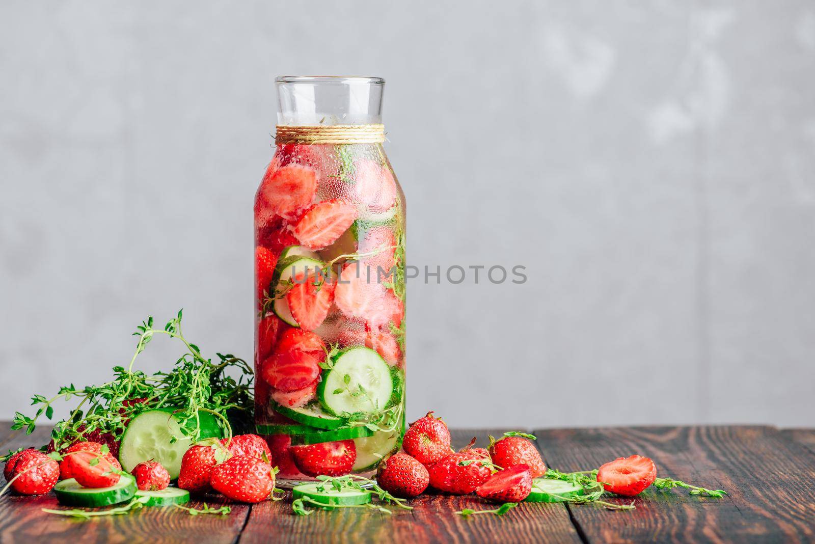 Bottle of Infused Water with Fresh Strawberry, Sliced Cucumber and Springs of Thyme. Ingredients Scattered on Wooden Table. Copy Space.