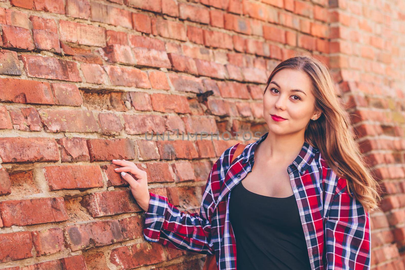 Portrait of young woman in checkered shirt and blue jeans standing against brick wall