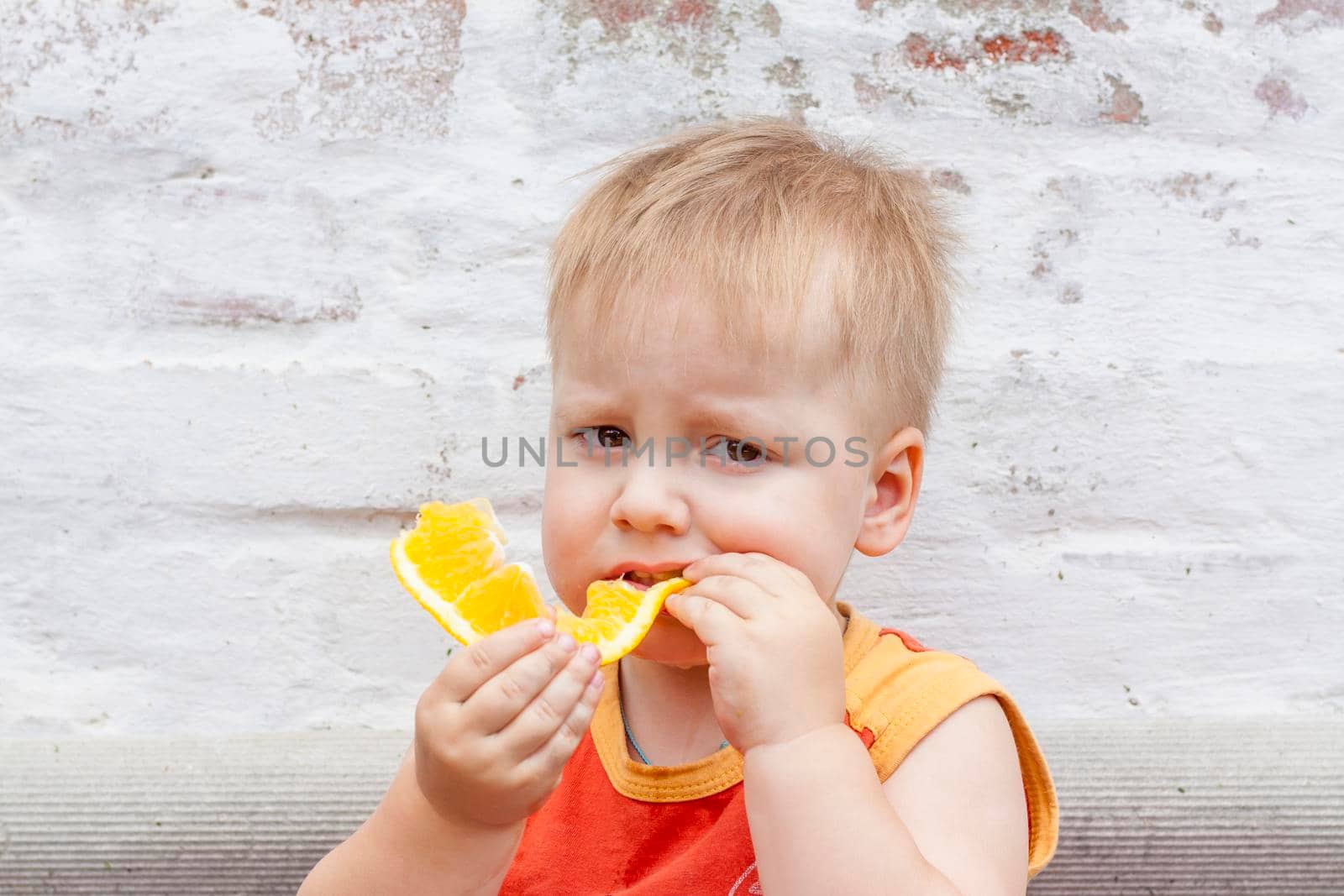 Portrait of child. Cute boy posing and eating a delicious orange. The emotions of a child. Portrait of child. Boy on the background of a brick wall. 