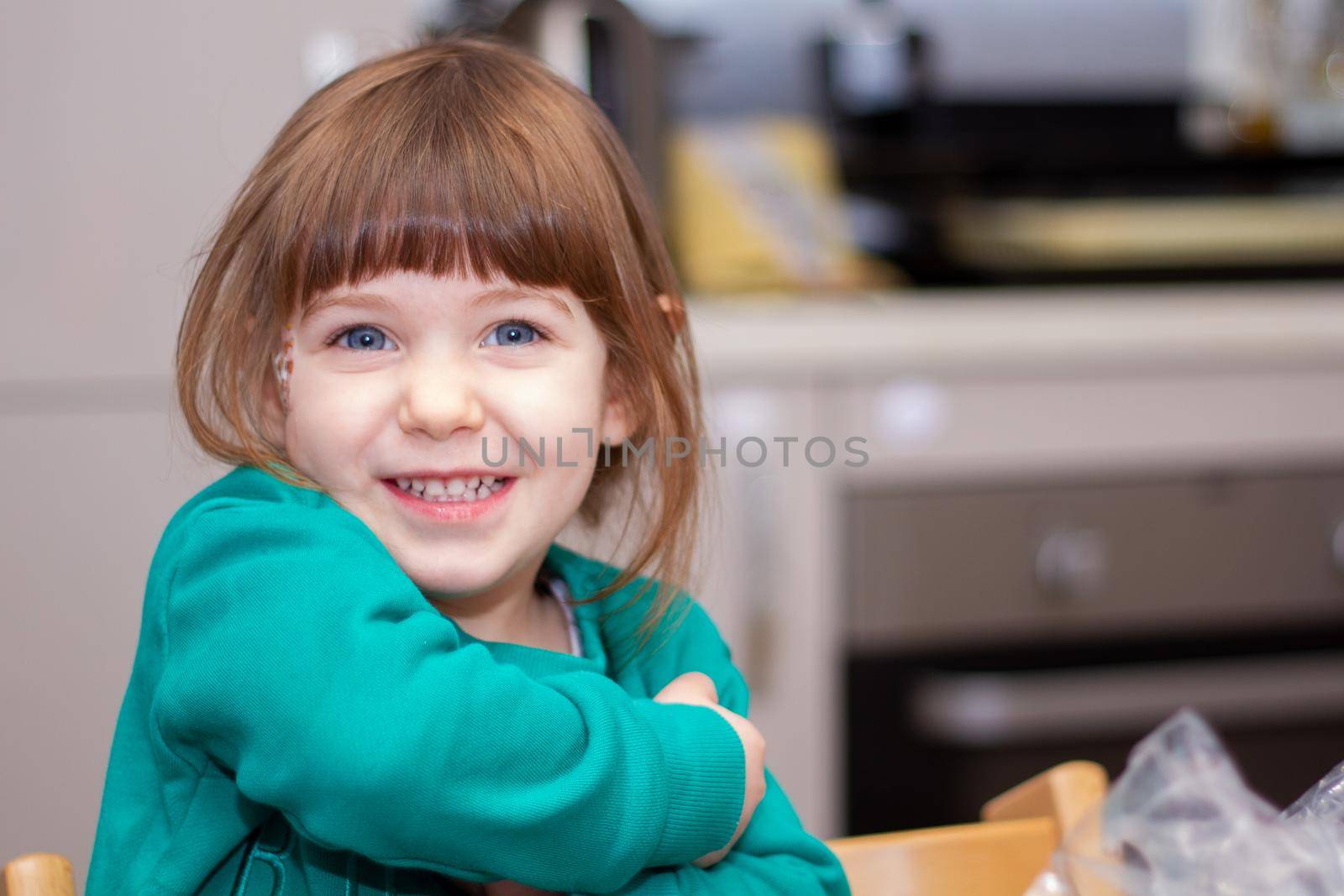 Portrait of a cute, brown-haired, blue-eyed, baby girl in green sweater in a kitchen