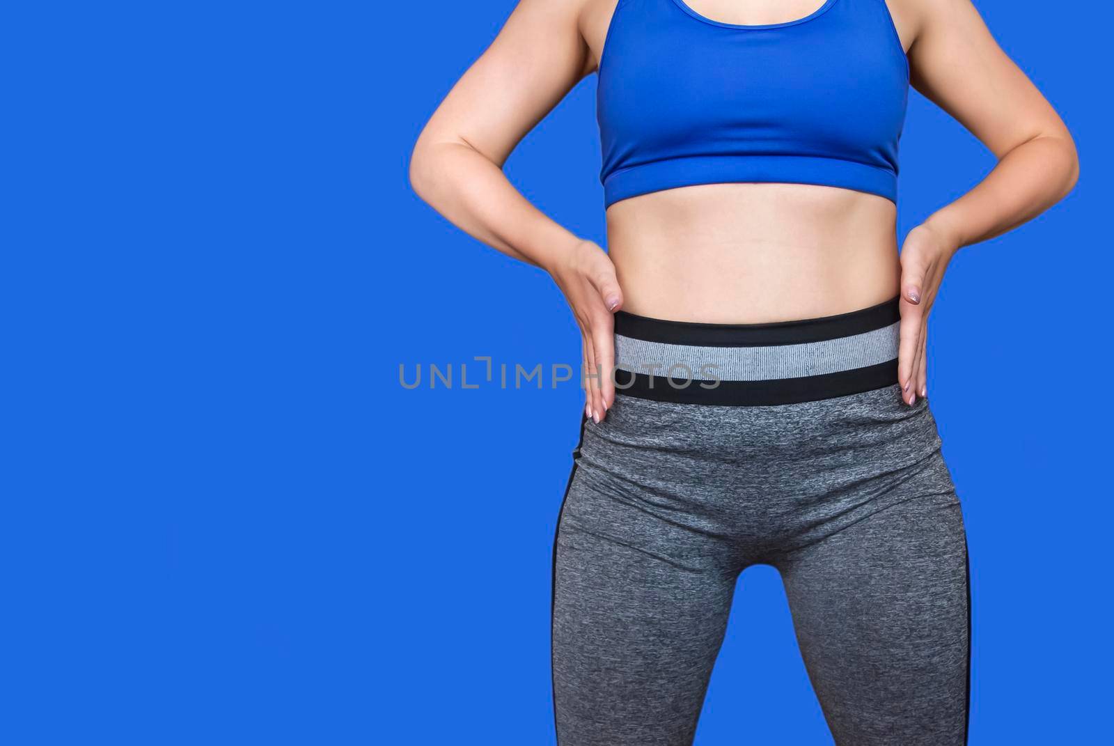 Fitness girl in a top and leggings points to her athletic figure. The Concept Of Weight Loss. Model on a blue background. A slender Young Lady Holds her hands at her waist. Free Space For Text.