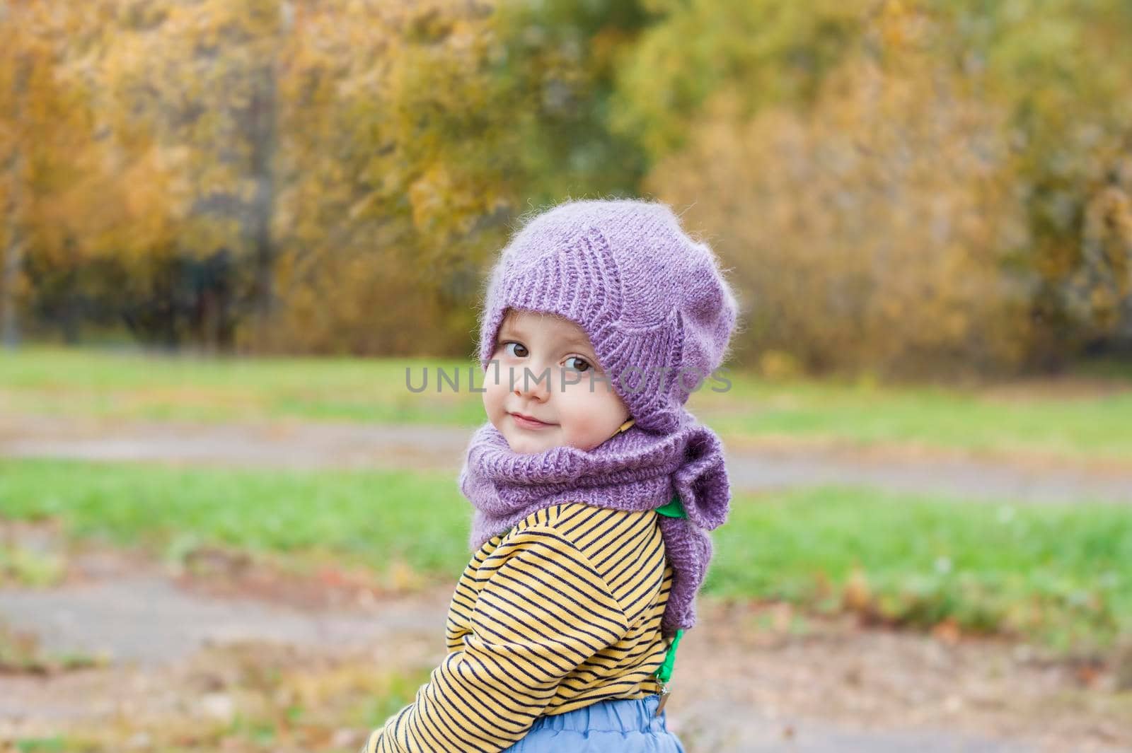 A child in a purple, knitted hat and scarf. Portrait of a child on the background of autumn trees. 