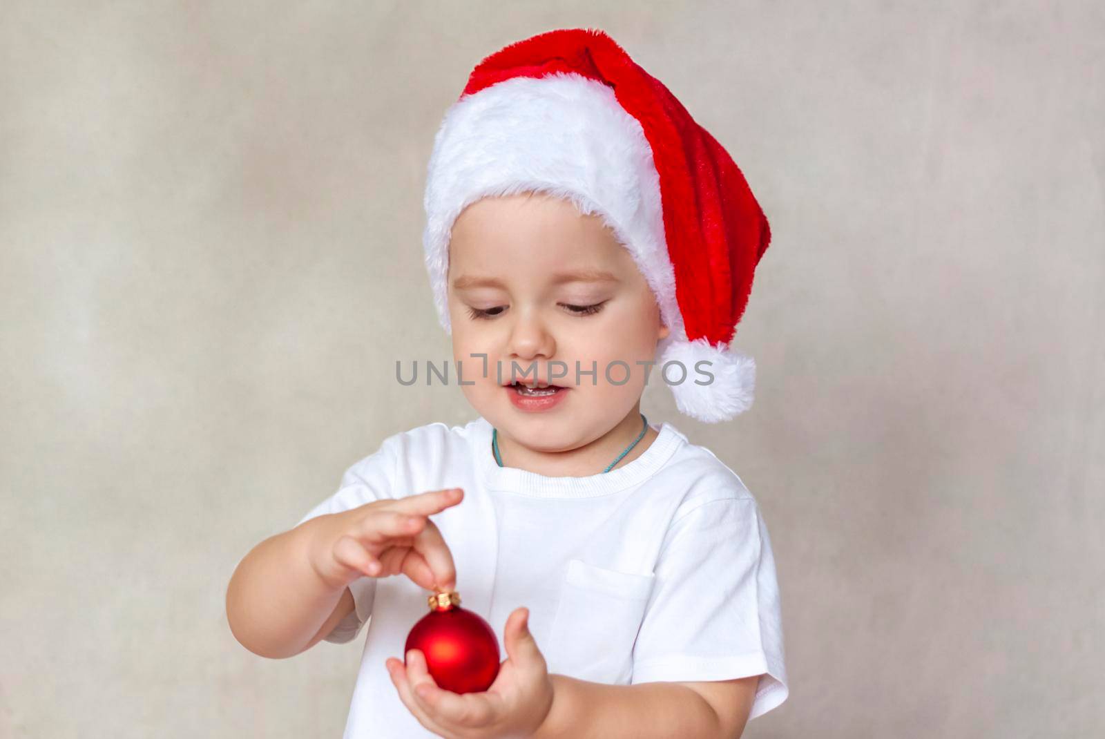 Portrait of a cute little boy in a white t-shirt and Santa Claus hat. A boy admires a red Christmas ball. Children's emotions. Christmas and new year. The concept of an advertising banner.