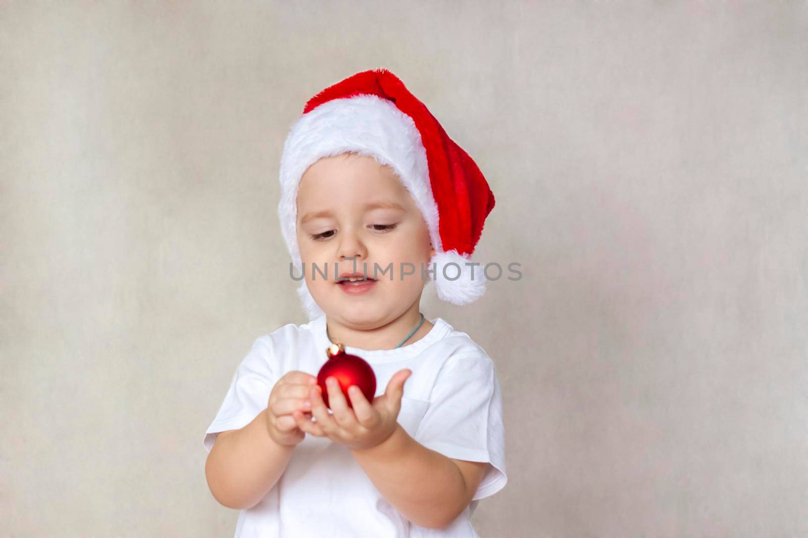 Portrait of a cute little boy in a white t-shirt and Santa Claus hat. A boy admires a red Christmas ball. Children's emotions. Christmas and new year. The concept of an advertising banner.  