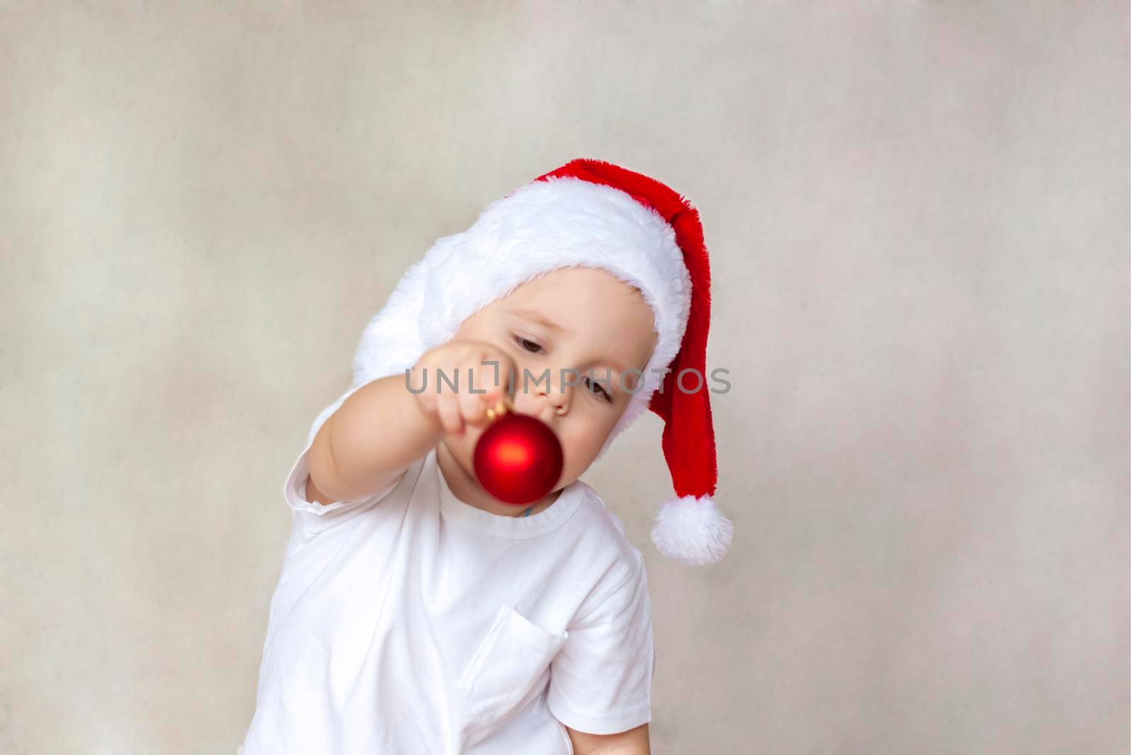 Portrait of a cute little boy in a white t-shirt and Santa Claus hat. A boy admires a red Christmas ball. Children's emotions. Christmas and new year. The concept of an advertising banner.