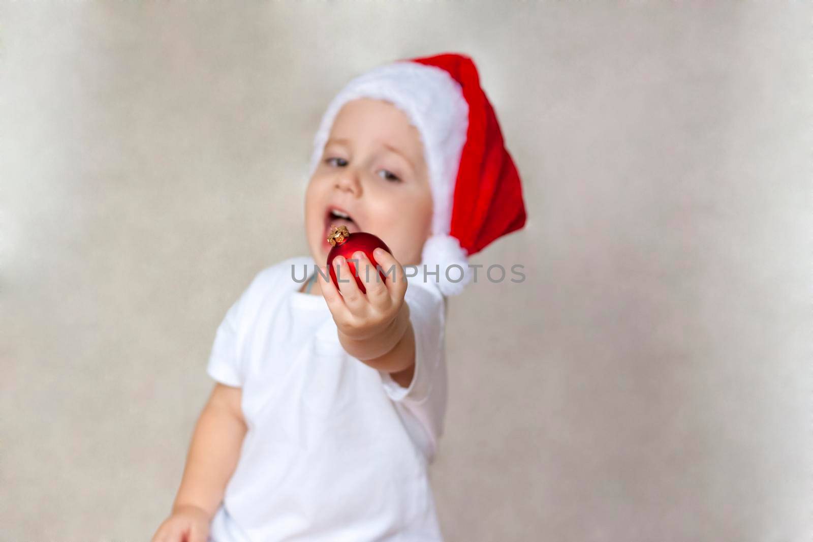 Portrait of a cute little boy in a white t-shirt and Santa Claus hat. A boy admires a red Christmas ball. Children's emotions. Christmas and new year. The concept of an advertising banner.