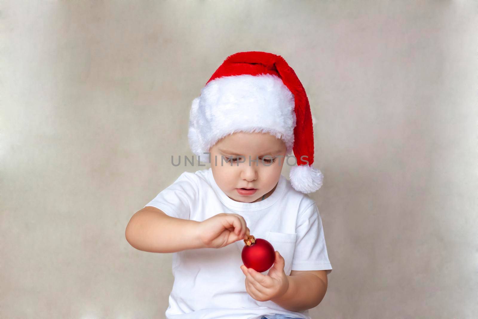 Portrait of a cute little boy in a white t-shirt and Santa Claus hat. A boy admires a red Christmas ball. Children's emotions. Christmas and new year. The concept of an advertising banner. 