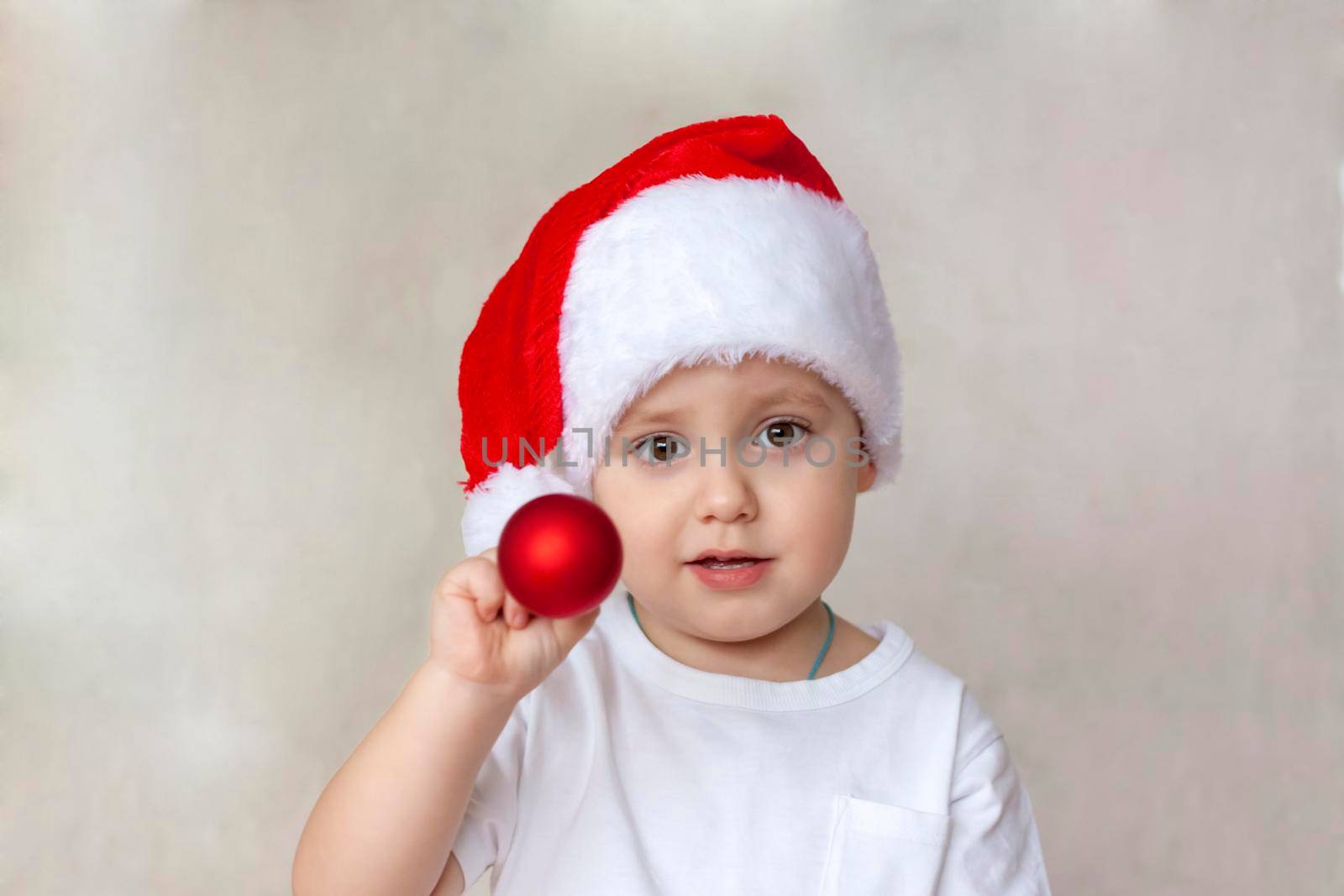 Portrait of a cute little boy in a white t-shirt and Santa Claus hat. A boy admires a red Christmas ball. Children's emotions. Christmas and new year. The concept of an advertising banner.
