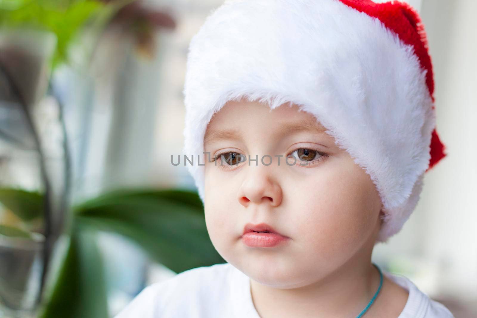 Portrait of a cute little boy in a white t-shirt and Santa Claus hat. Children's emotions. Christmas and new year. The child looks out the window. The concept of an advertising banner.