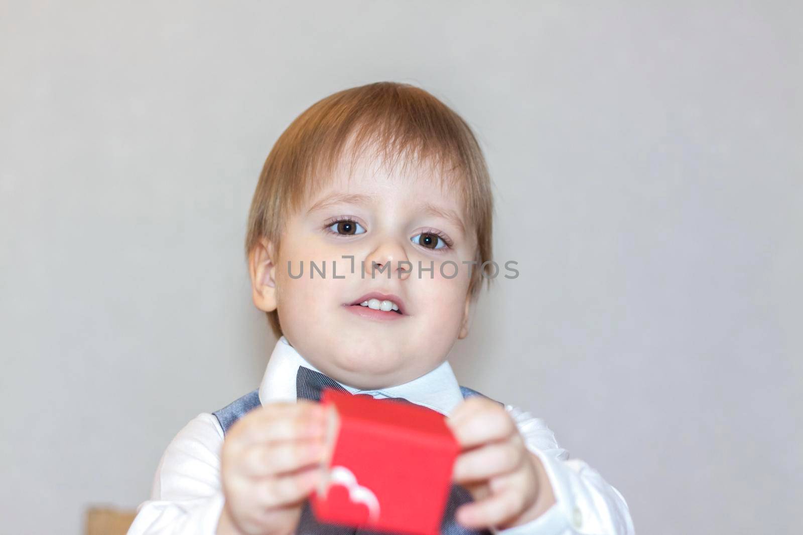 A little boy holds and hands over a red box, a Valentine's Day theme concept. Portrait of a cute boy in a suit with a bow tie. Valentine's Day. 