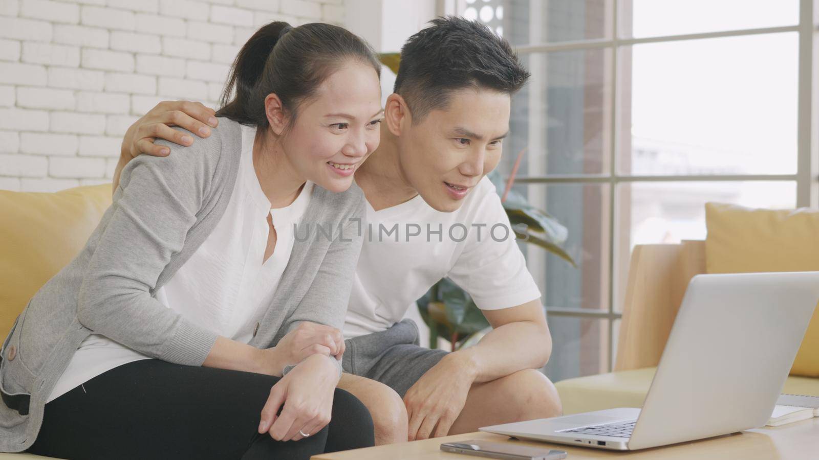 Happy Asian family couple husband and wife laughing sitting on sofa using laptop computer webcam technology talking making online social distance video call to their children at home.