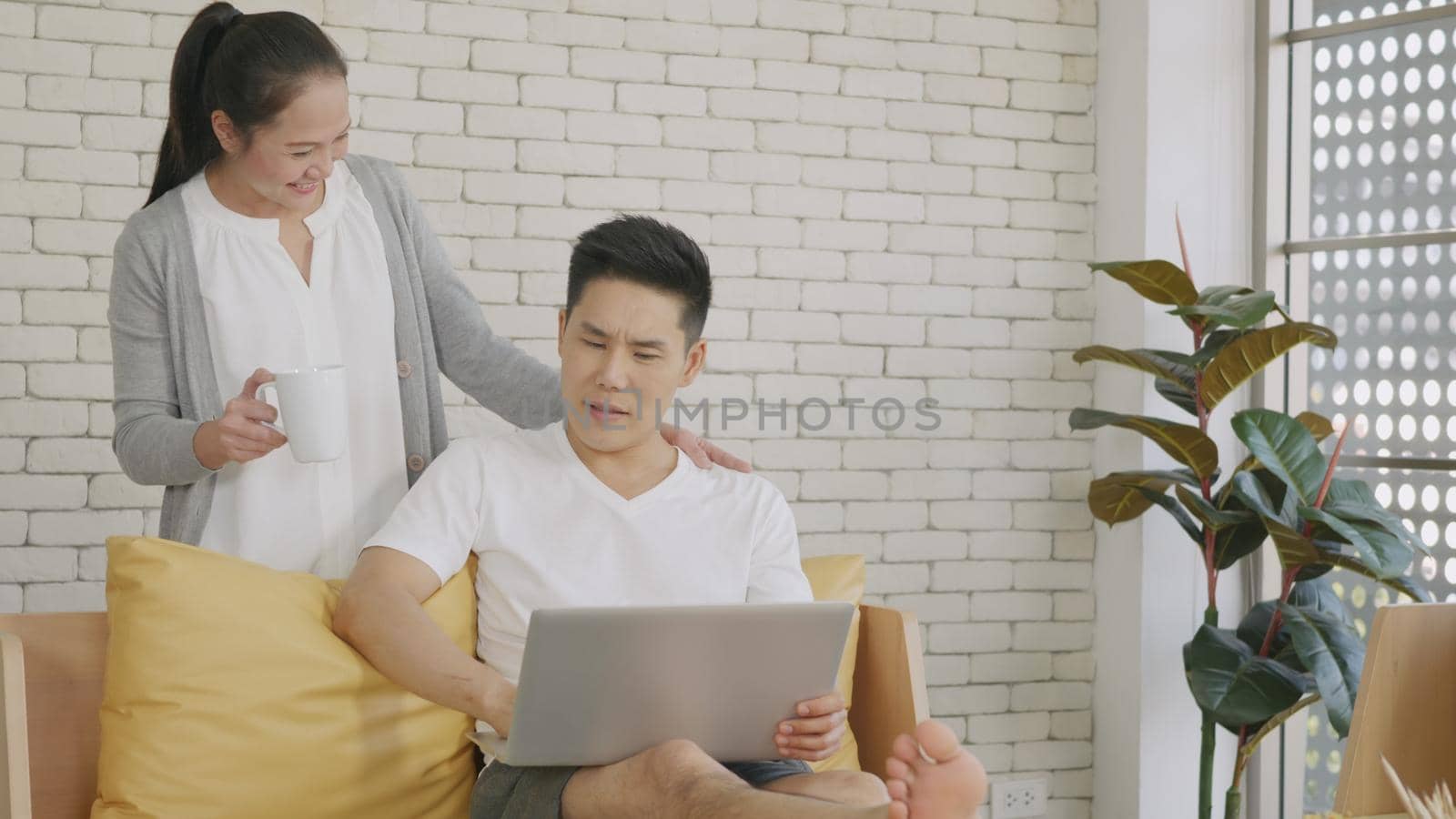 Happy Asian beautiful family couple husband and wife laughing sitting on sofa in the living room working with laptop computer at home. Woman brings coffee to the man during work