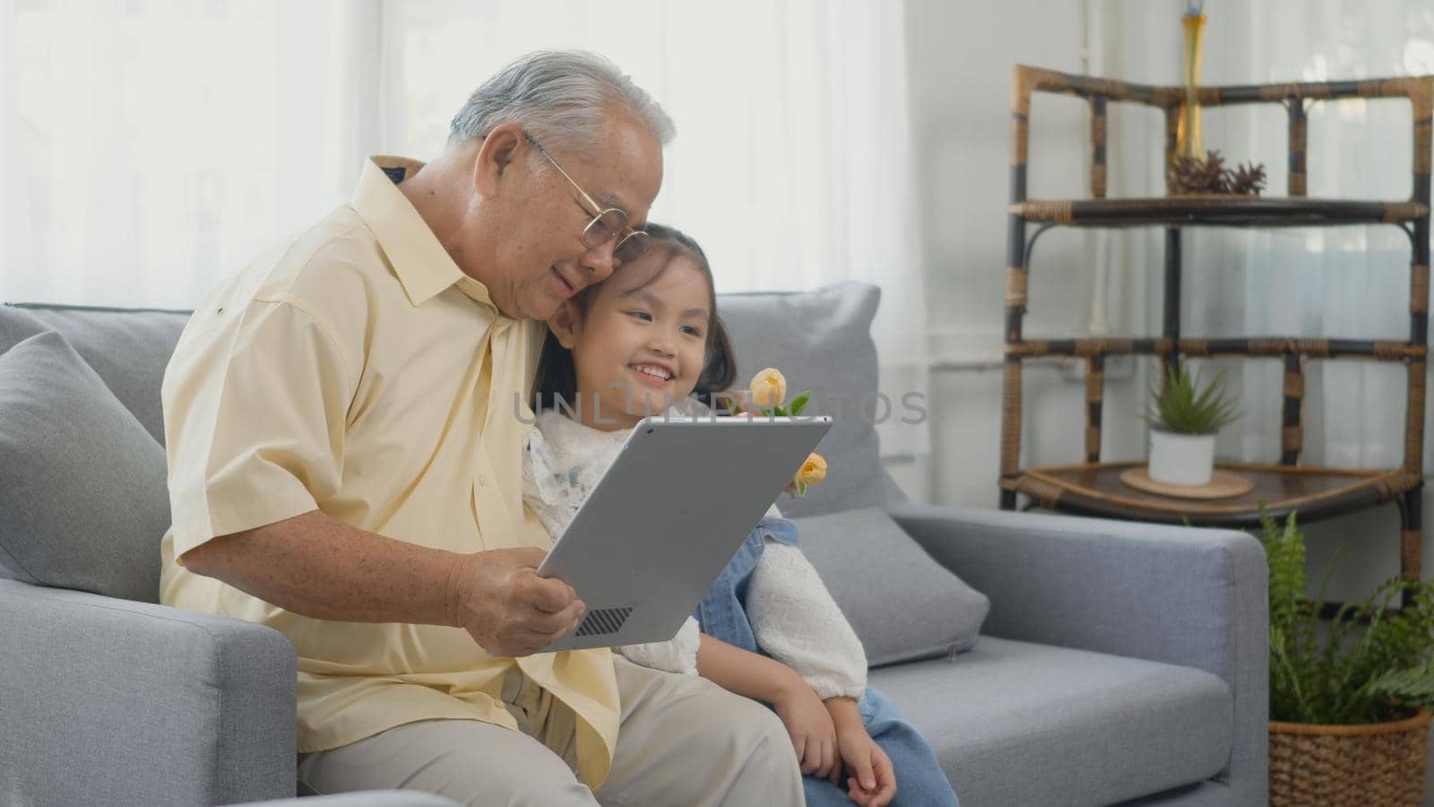 Asian senior old man looking to tablet computer and granddaughter come visitor at home, Grandfather reading news on digital tablet with his kid on sofa in living room