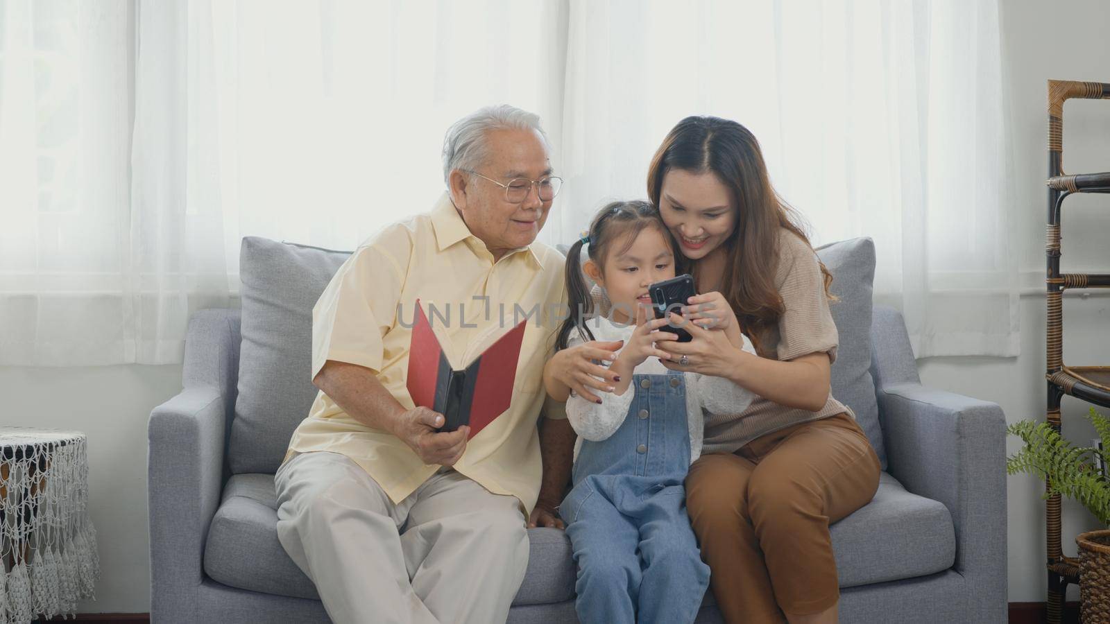 Asian grandparents laughing taking selfie with granddaughter on sofa at home by mobile by Sorapop