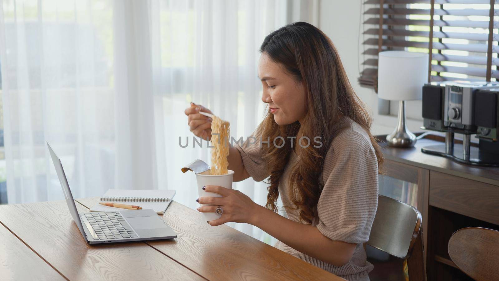 Asian business woman eating instant noodles while working on laptop computer at home office, Happy beautiful young female sitting on desk work overtime doing deadline project, late time business