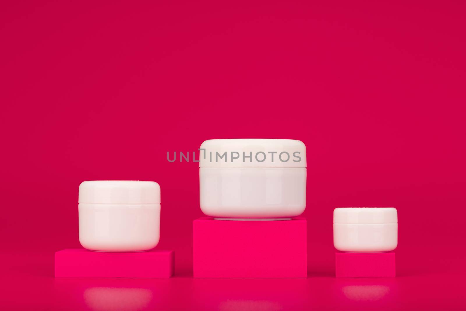 Set of creams for face, neck and under eye skin on pink pedestals against pink background with copy space. Unbranded cosmetic jars on shopfront. Concept of skin care and beauty