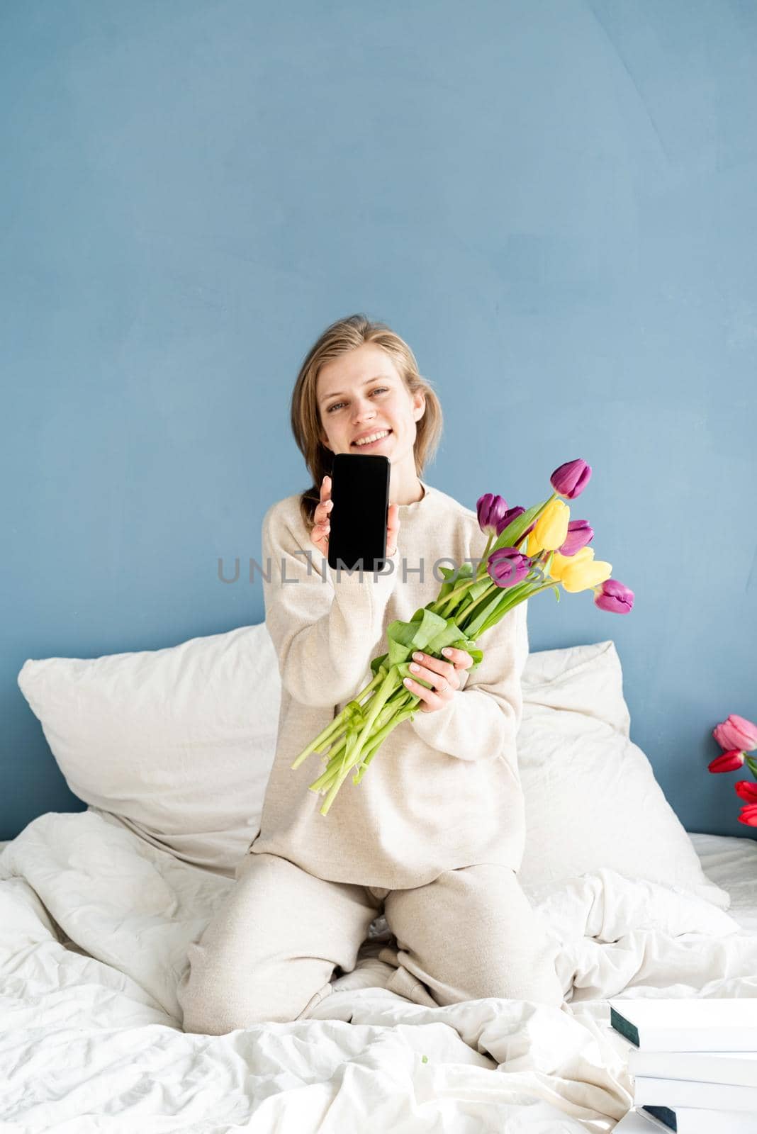 Happy woman sitting on the bed wearing pajamas holding tulip flowers and showing smartphone screen, mock up design