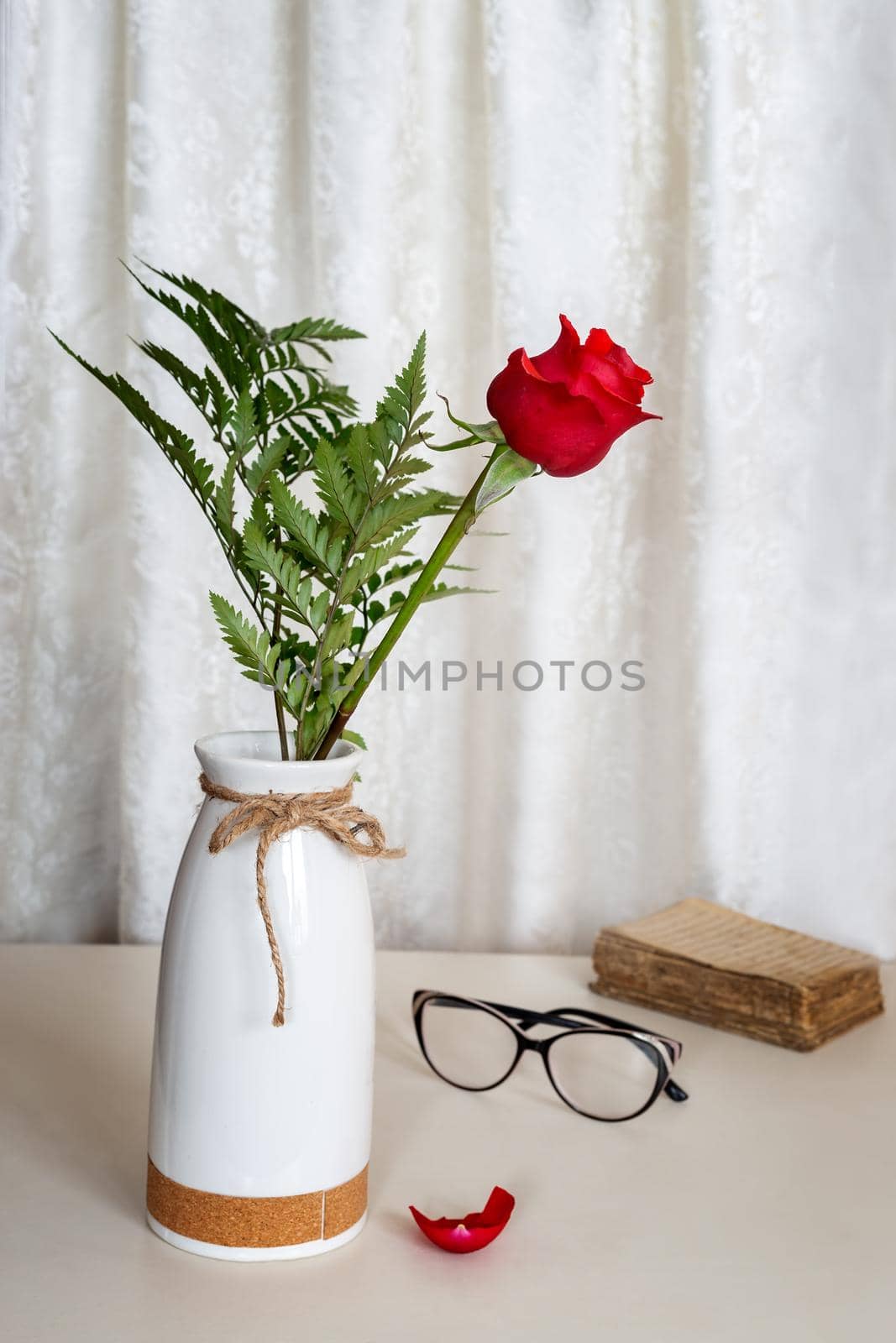 On the table in a white ceramic vase is a flower of a beautiful red rose. Next to an old book and glasses. Front view, copy space