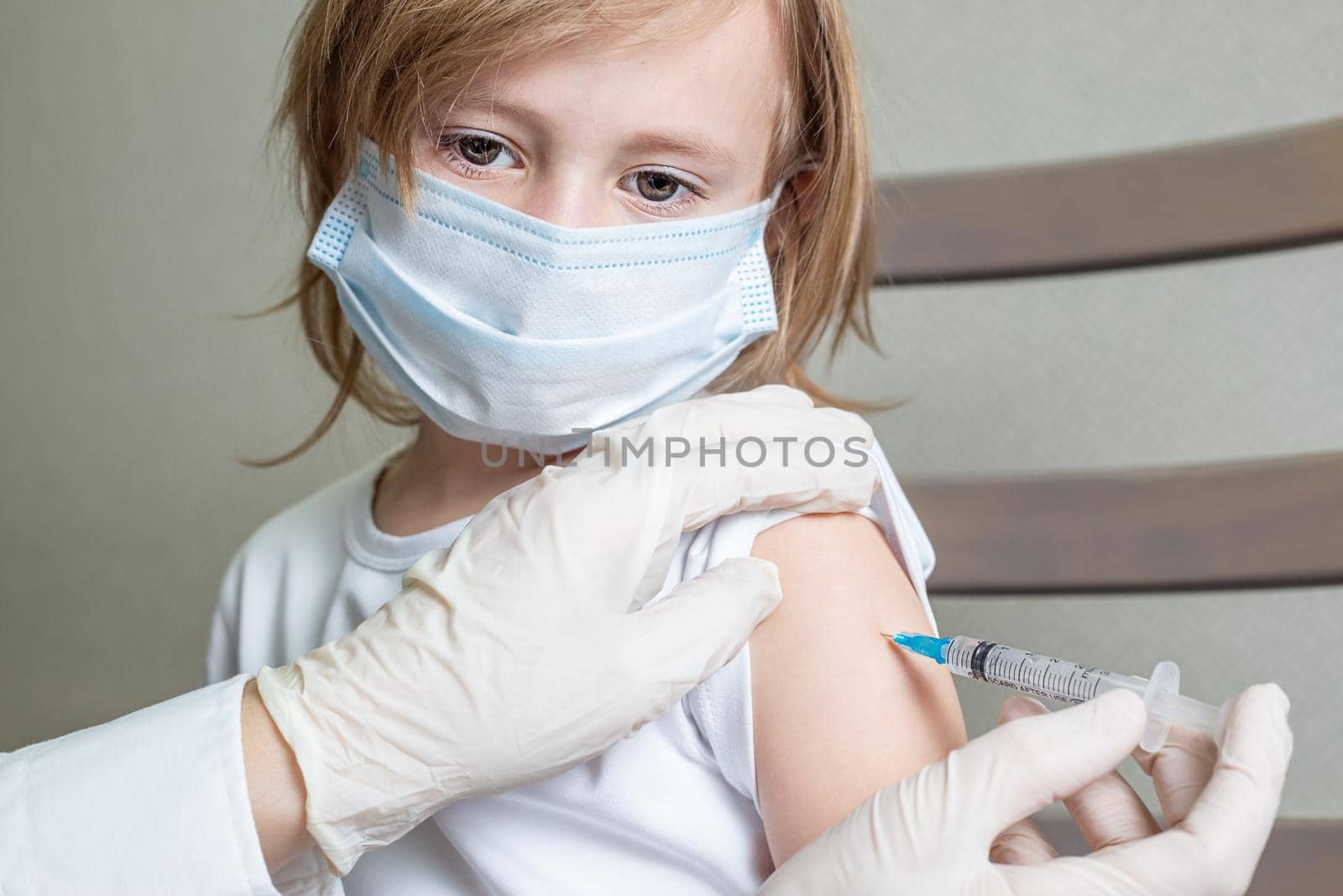 Doctor in a medical mask and gloves makes an injection in the shoulder of a little caucasian girl in a medical mask, the girl looks aside