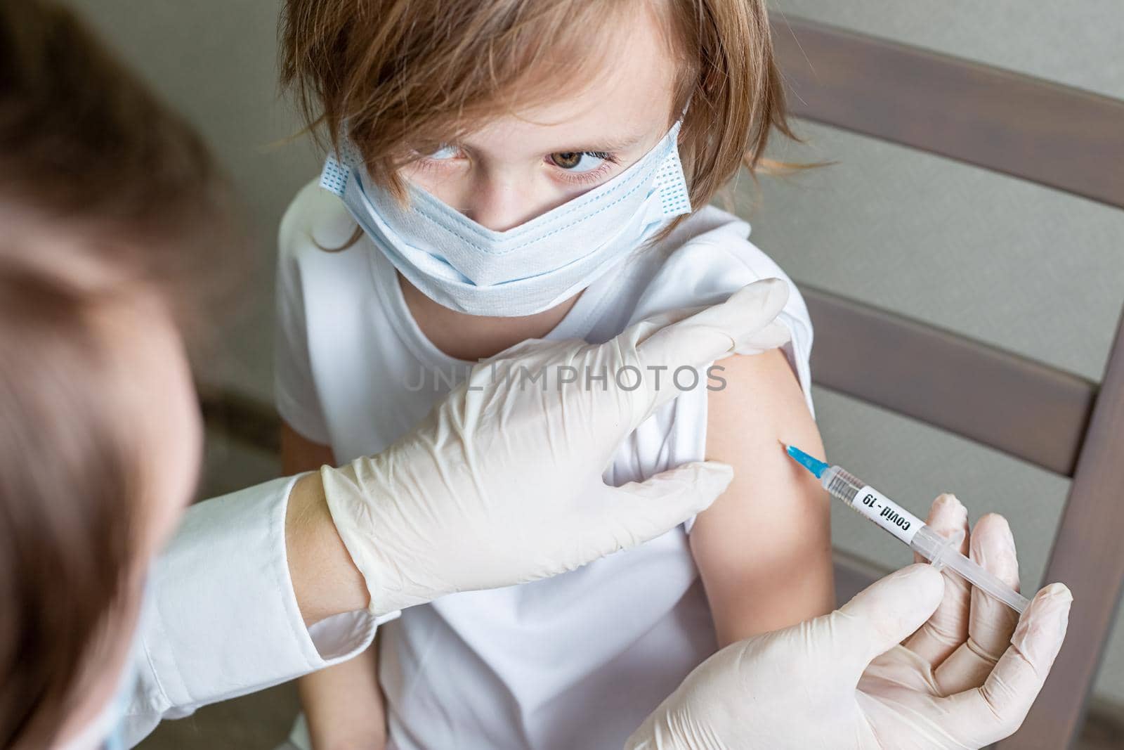 Doctor in a medical mask and gloves makes an injection of a vaccine against a coronavirus infection into the shoulder of a little caucasian girl in a medical mask, the girl looks at the doctor
