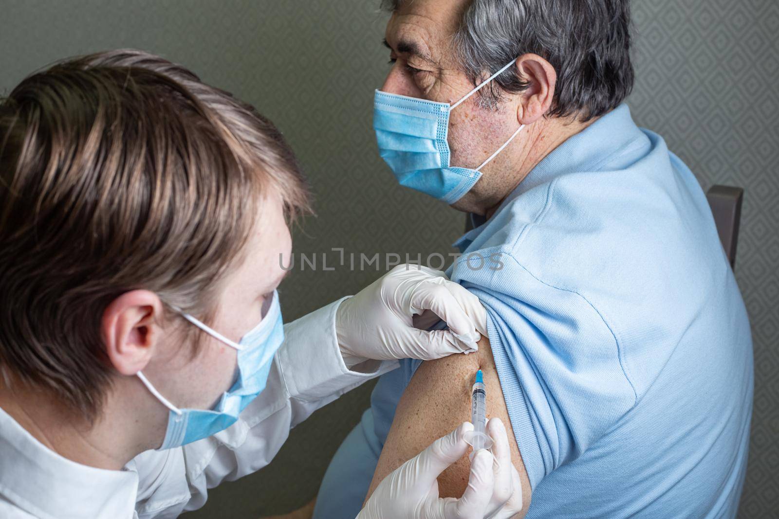 An elderly man of Caucasian appearance in a mask receives a dose of coronavirus vaccination at home, the doctor came to the patient to give a protective injection against the disease
