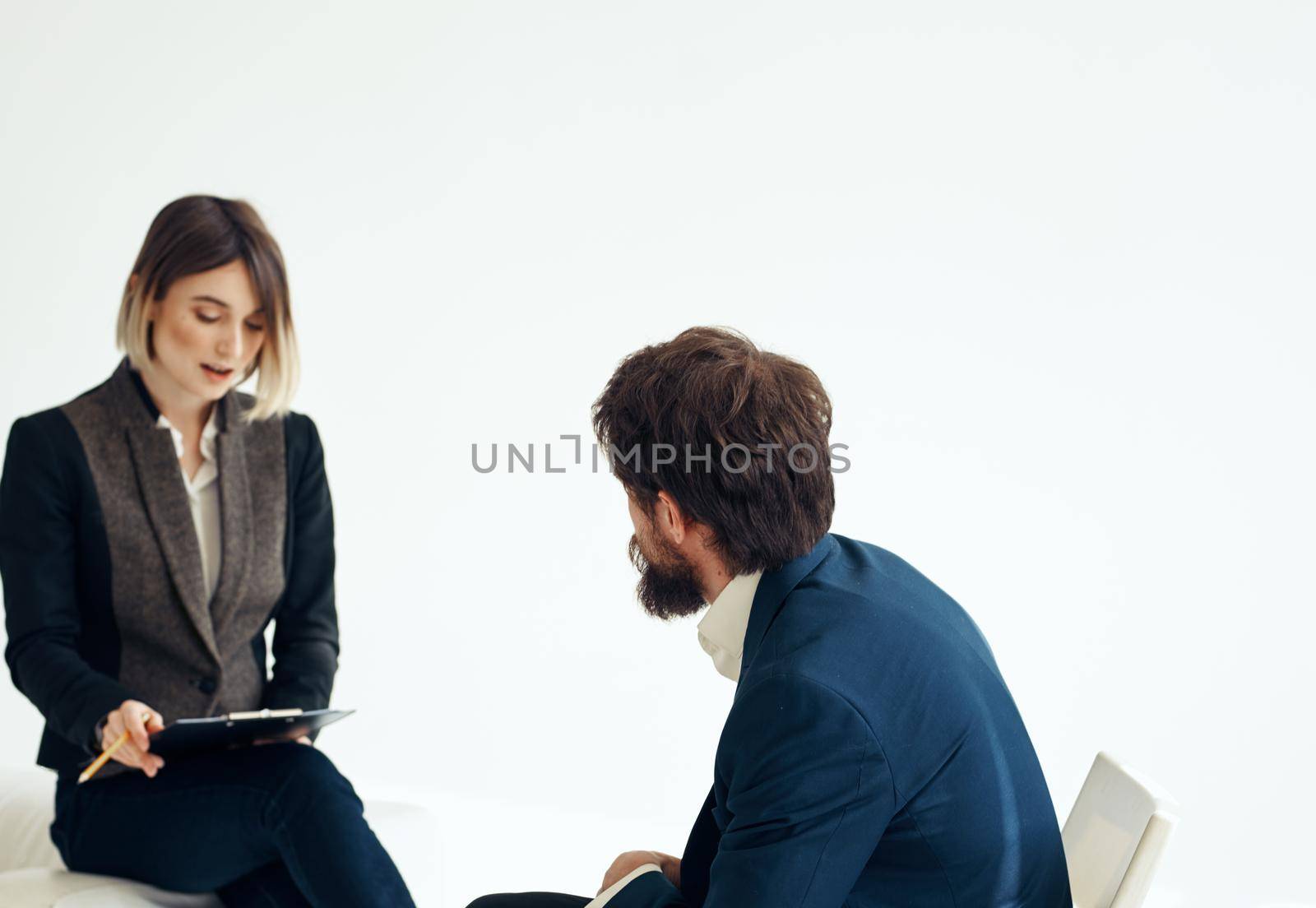 A man in a blue jacket sits opposite a woman in a suit on a light background indoors. High quality photo