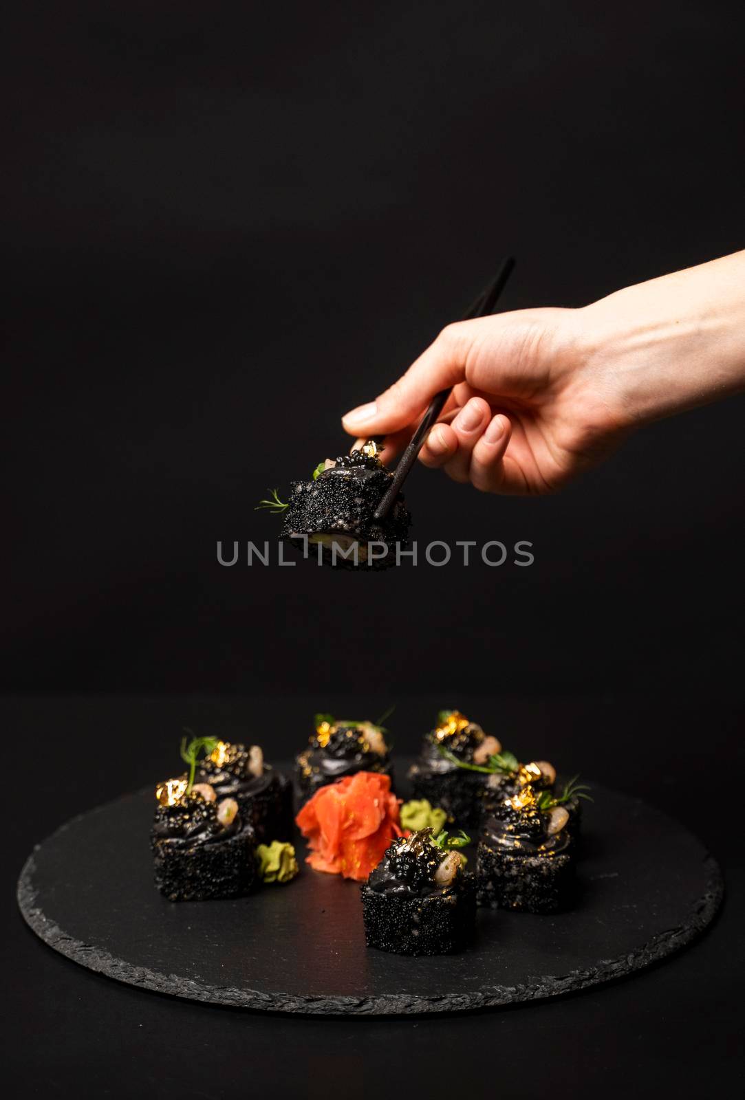 Hand with chopsticks holds a sushi. Custom sushi roll with black rice, crab meat, avocado, smoked salmon mousse, oar caviar, masago, shrimp cocktail, edible gold leaf, ginger, wasabi on black table