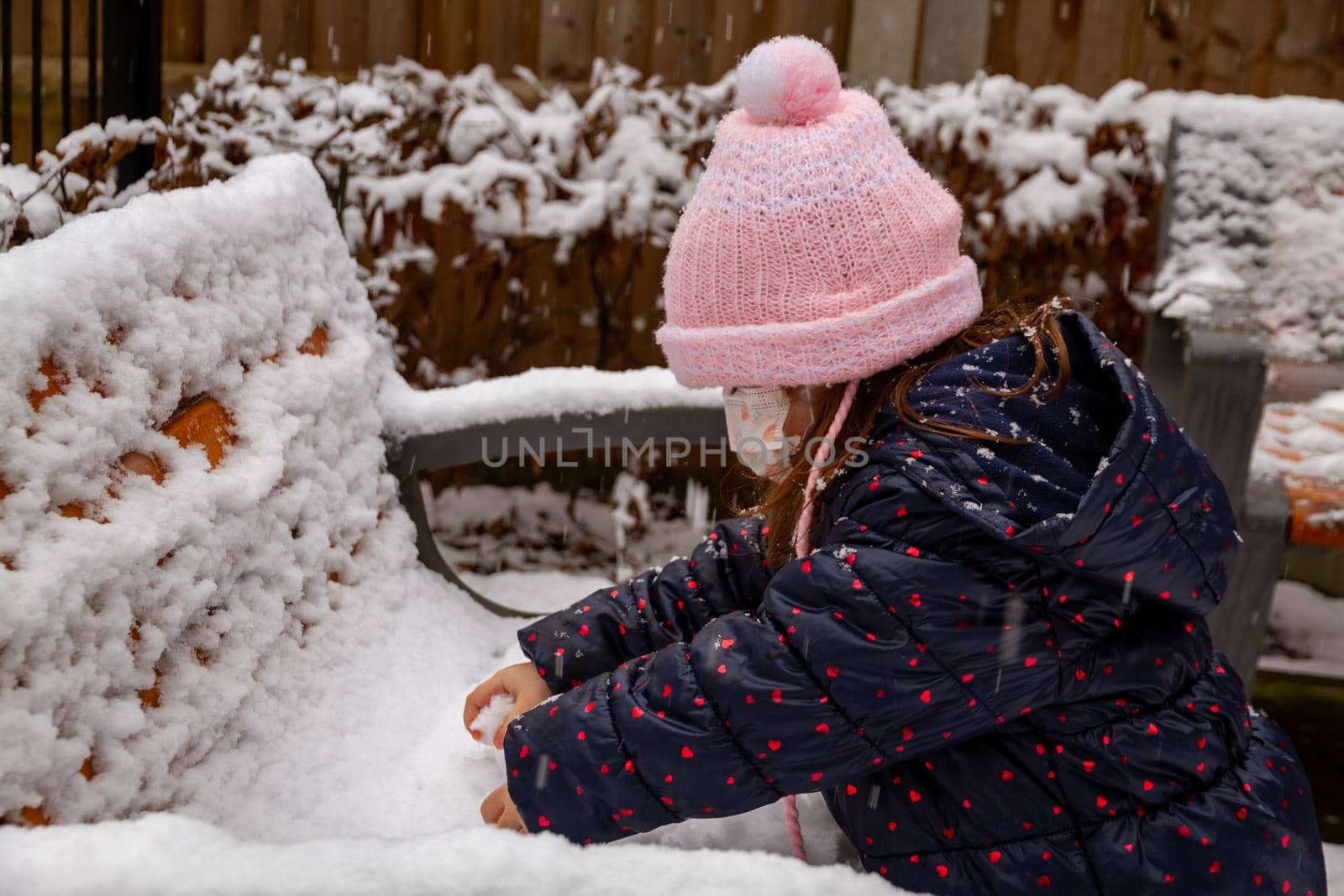 Portrait of a cute baby girl in pink hat playing in the snow wearing a face mask