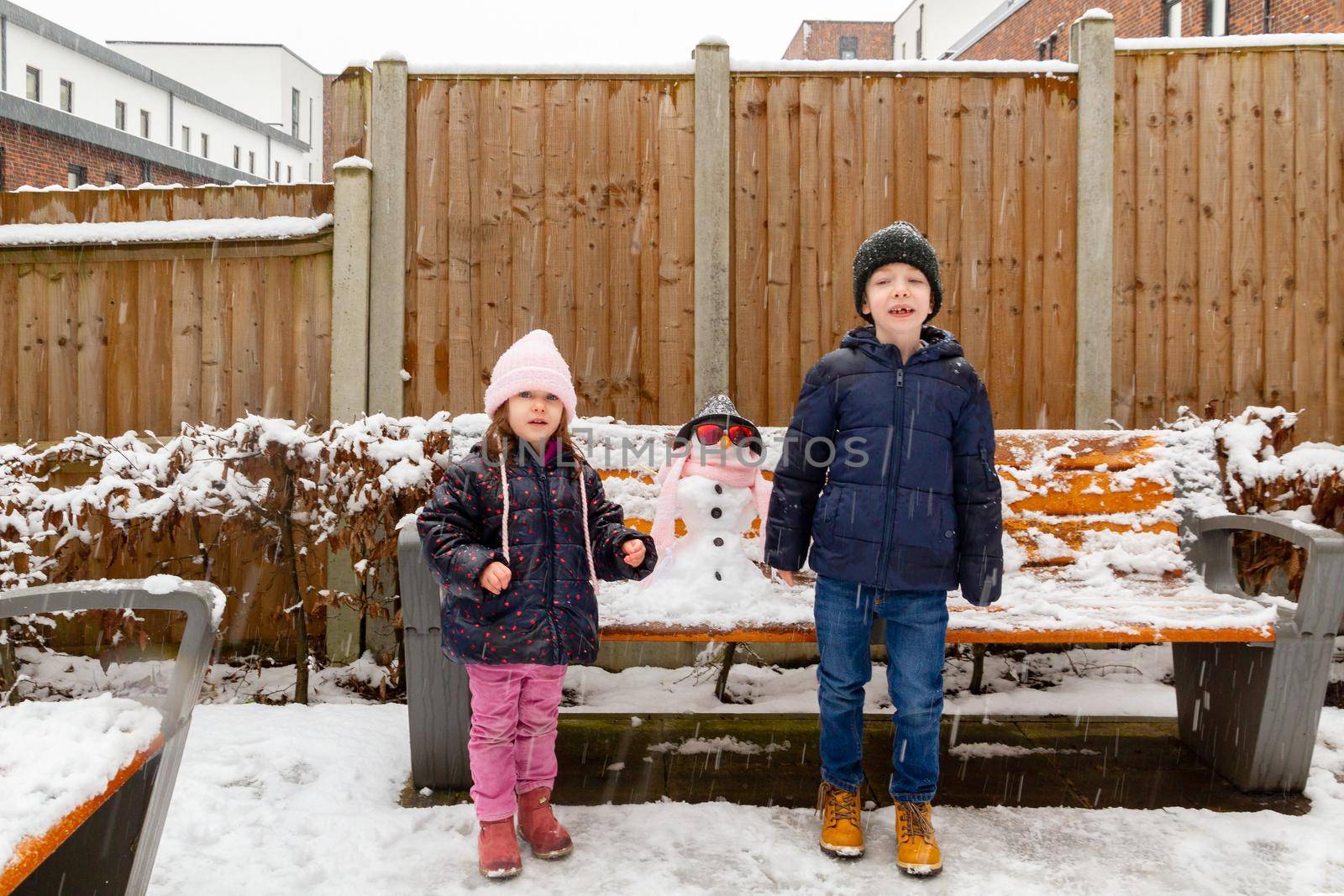 A cute blue-eyed, redhead boy and a cute, blue-eyed, brown-haired girl standing in the snow by a snowman