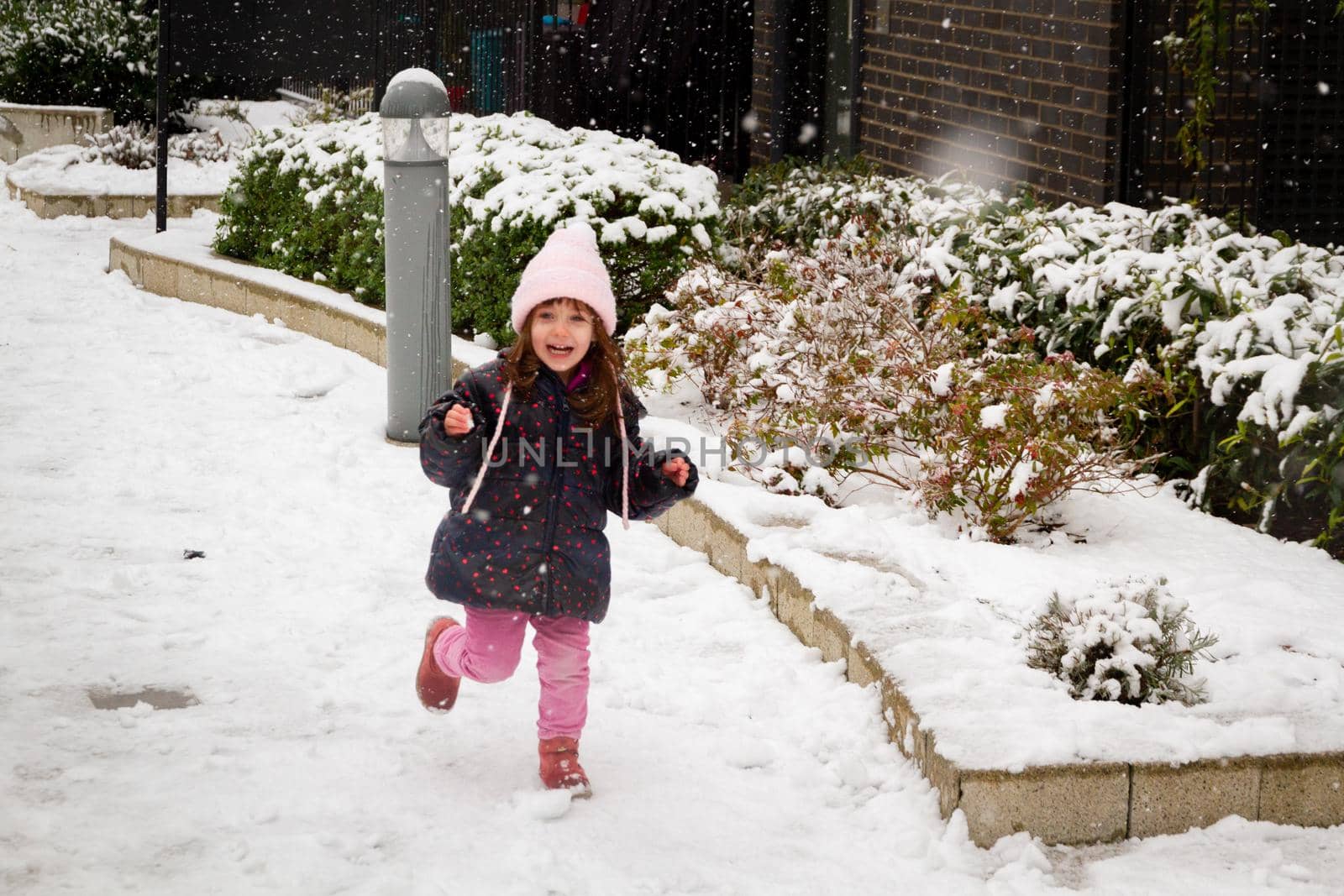 A cute, blue-eyed, brown-haired girl wearing a pink hat and ablue coat running in the snow
