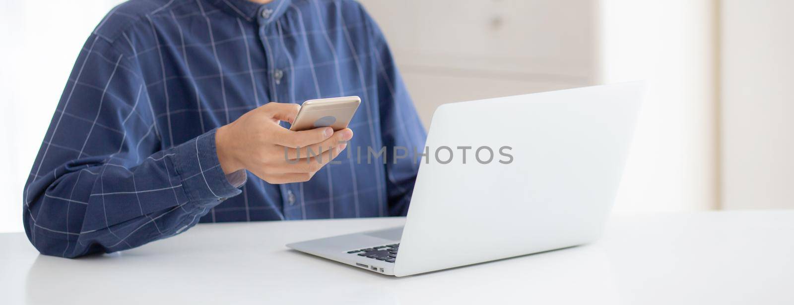 Closeup hand of young man working laptop computer and reading smartphone on internet online on desk at home, freelance male using phone with social media, business and communication concept. by nnudoo