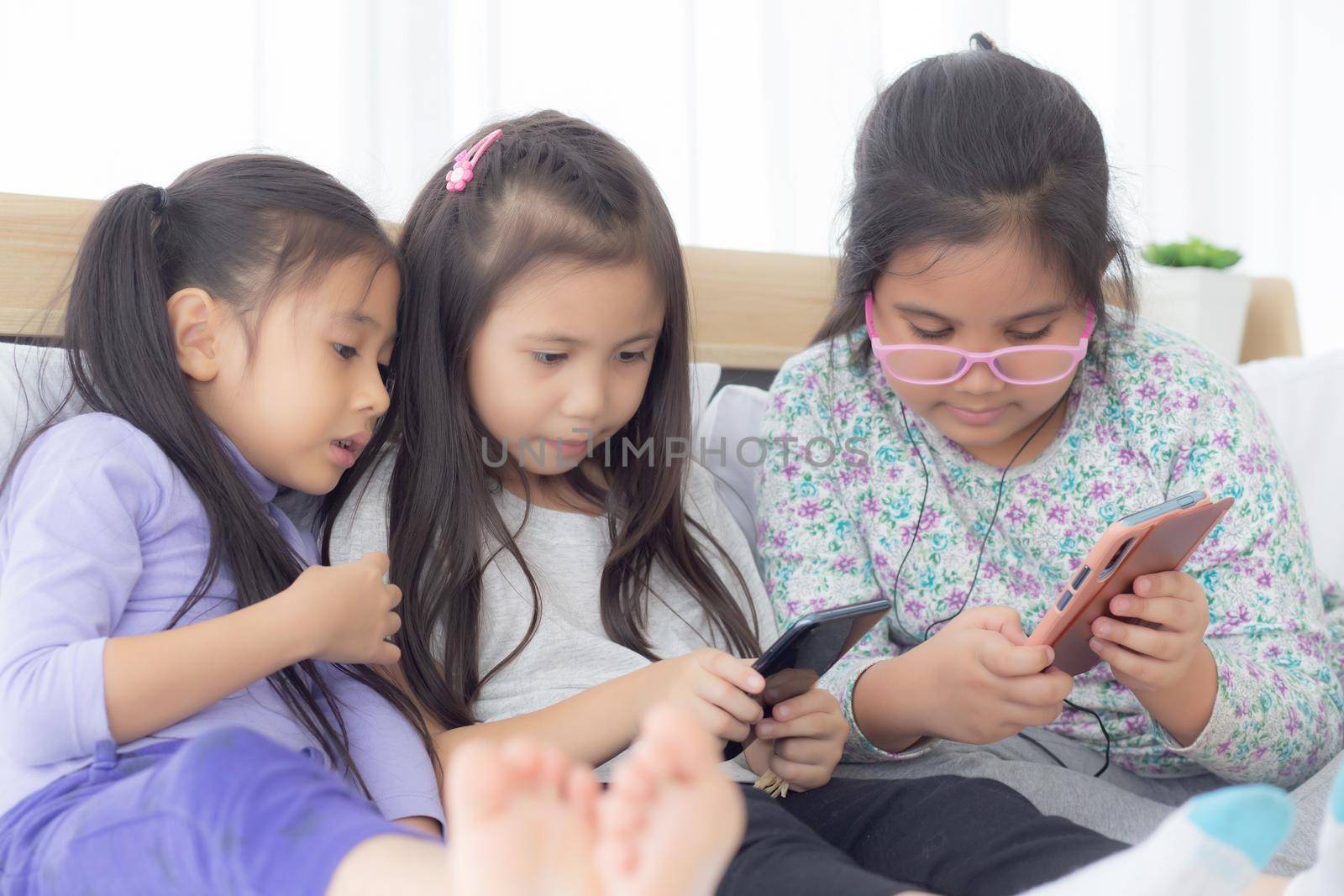 Happy asian little kid and friends playing smartphone on sofa at home, children using phone together on couch, girl watching smart phone for entertainment, lifestyle and communication concept.