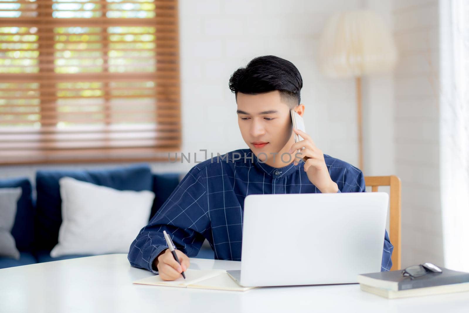Young asian man talking phone and work from home with laptop computer, freelance male writing on notebook and speak on smartphone, stay home, domestic life, business and communication concept.