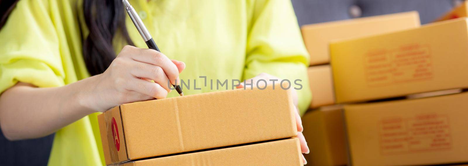 Closeup hand of young asian woman writing address on parcel box at home for delivery order to customer, shipping and logistic, merchant online and seller, business owner or SME, online shopping.