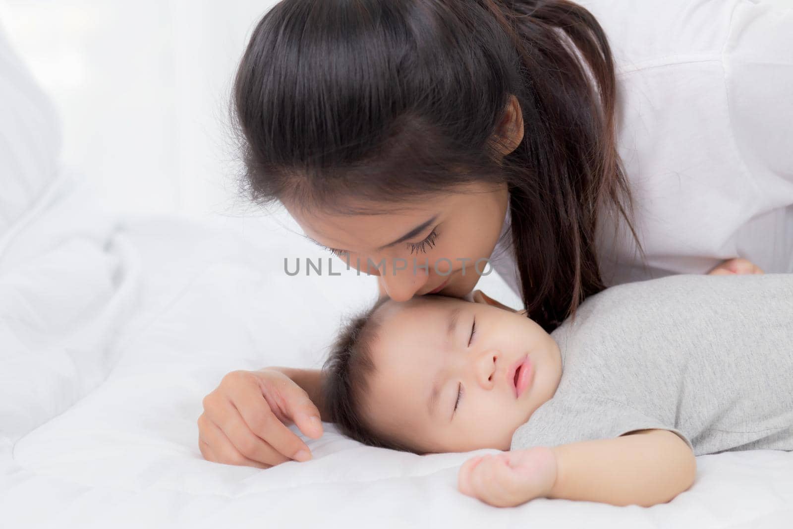 Young asian mother kiss cheek of little baby girl with tender on bed in the bedroom, mom love newborn and care, mother with expression with child together, parent and daughter, family concept.