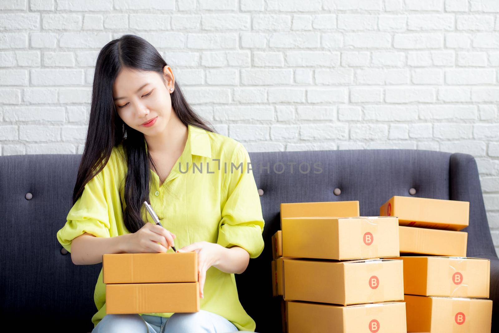 Young asian woman writing address on parcel box at home for delivery order to customer, shipping and logistic, merchant online and seller, business owner or SME, online shopping and e-commerce.