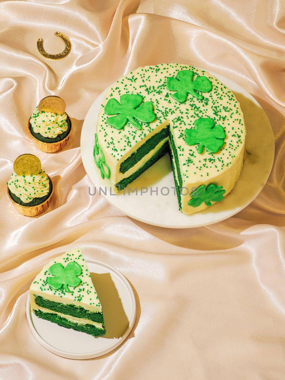 St Patricks Day sweet food concept. Sweet desserts for Saint Patrick's Day party - cake decorated shamrocks, green velvet cupckakes, chocolate golden coins and horseshoe on satin. Copy space. Vertical